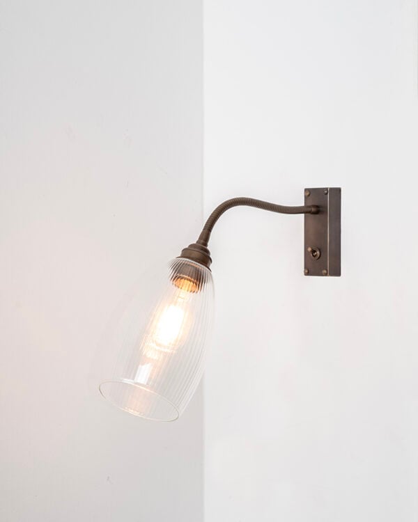 Antique Brass metal finish flexible wall light installed on white wall, featuring skinny ribbed glass Upton glass shade