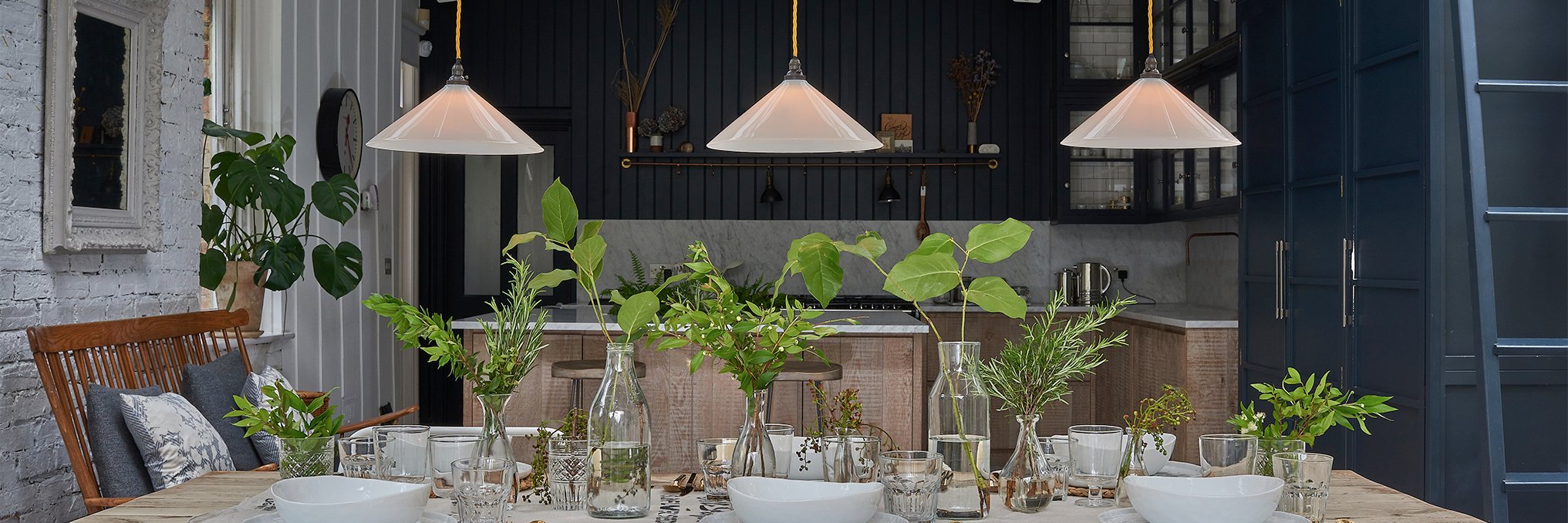 Three white Hay pendant lights suspended over a kitchen island. Decorated with multiple vases of plants, part of the Hay lighting collection.