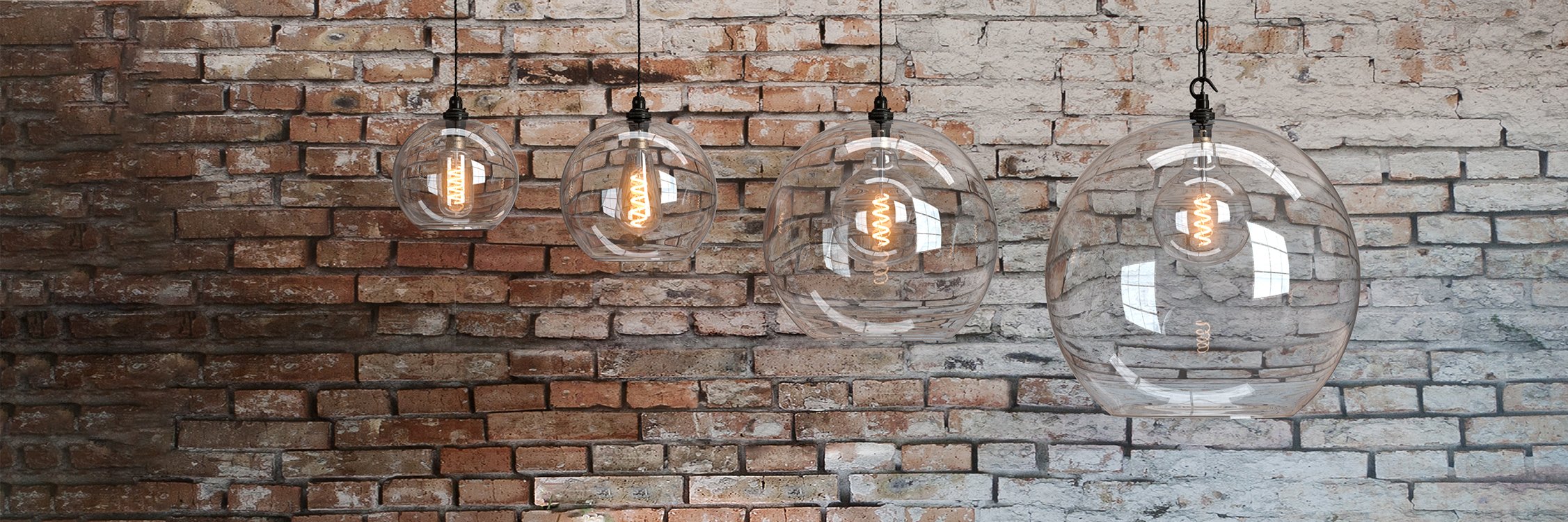 Four clear glass pendant lights hung against a brick wall. All in a different sizes, part of the Hereford lighting collection.