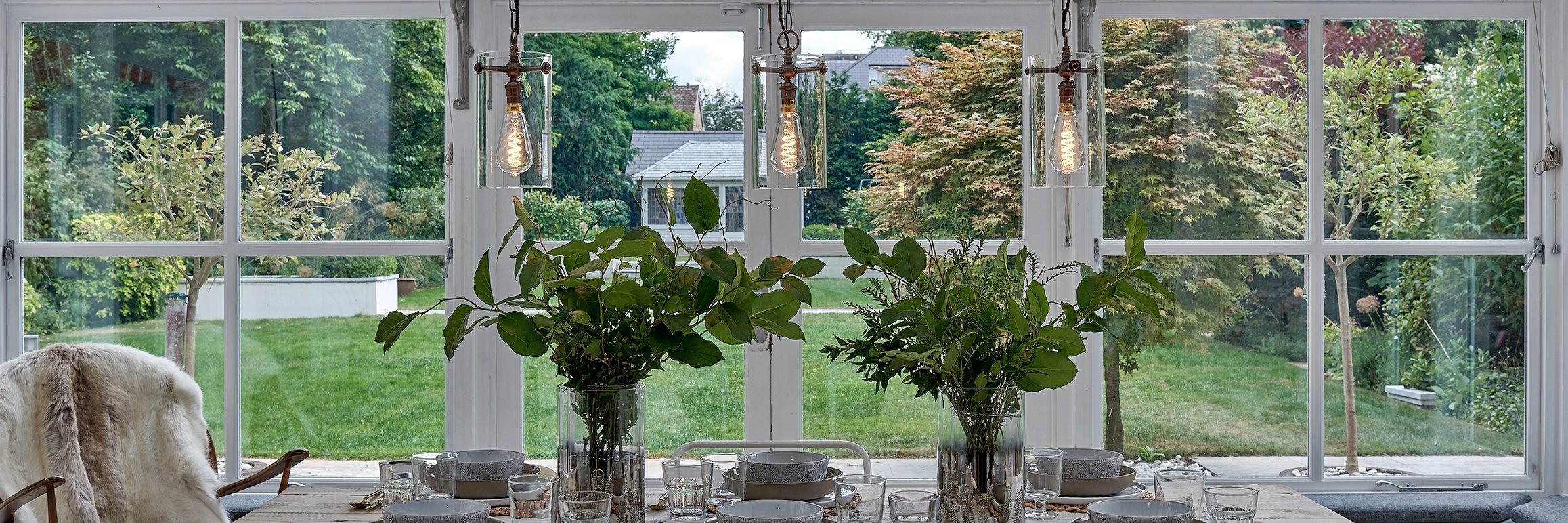 A green dining area with three clear glass pendant lights from the Sellack lighting collection, hanging above the table which is decorated with plants.